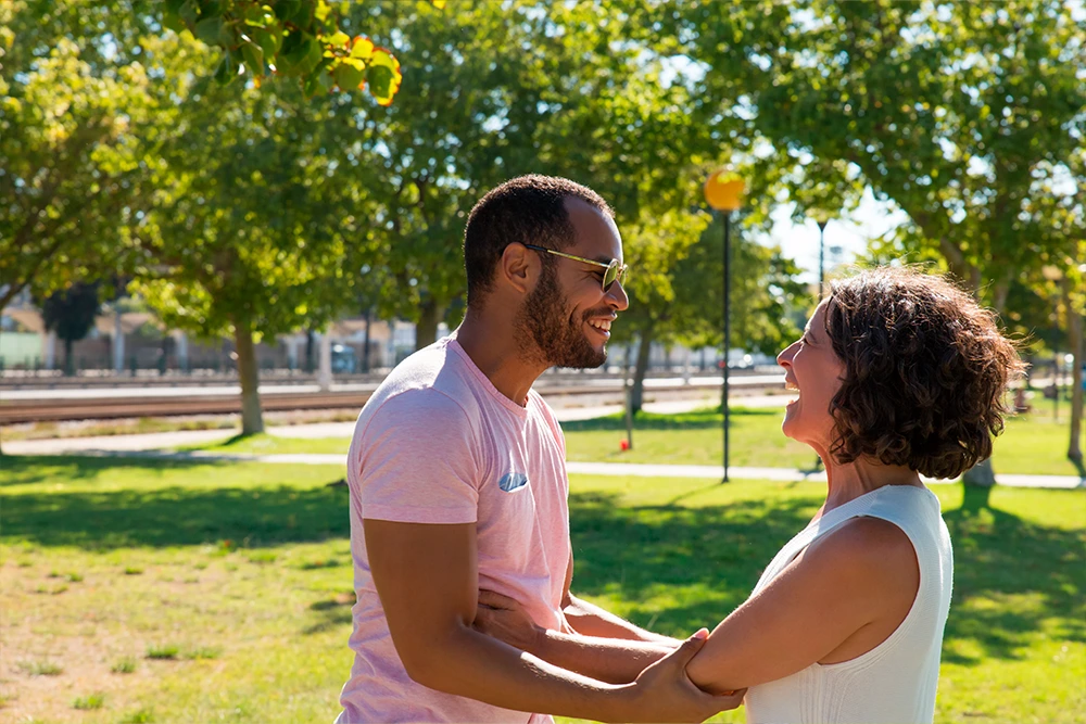 Multi-ethnic man and woman smiling at each other while holding hands in a sunny park.