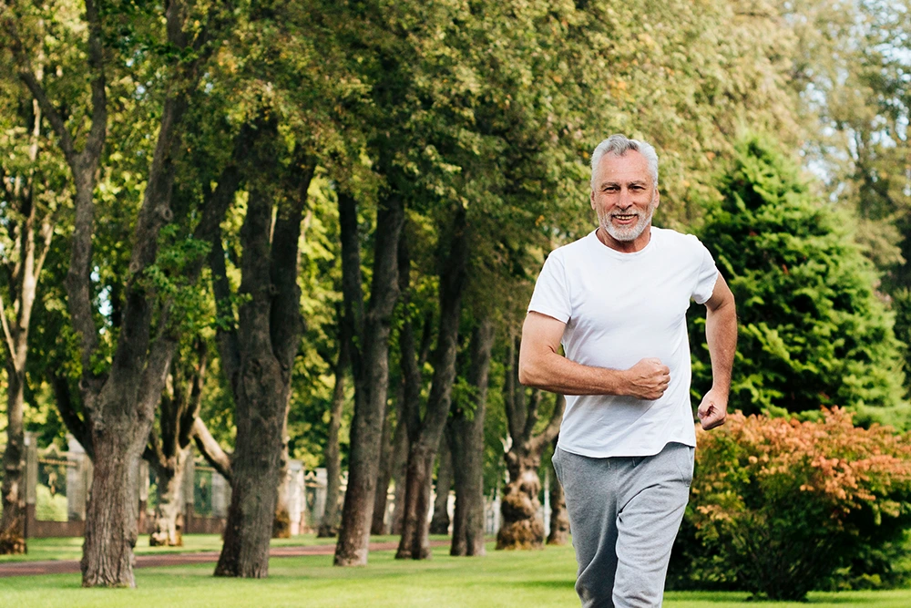 Older man with grey hair and beard jogging on a path in a wooded park.