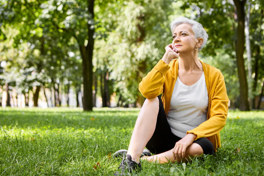 Mature woman with grey hair sitting on the grass in a park, looking thoughtful.